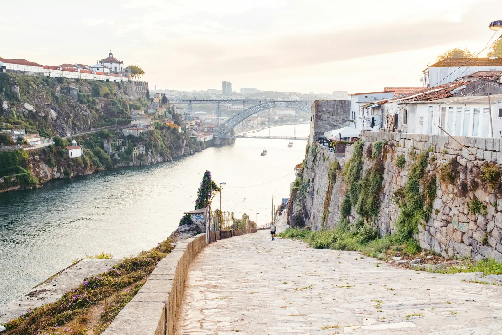 A quiet stone walkway along the Douro river in Porto, Portugal, with the Dom Luis I bridge in the distance