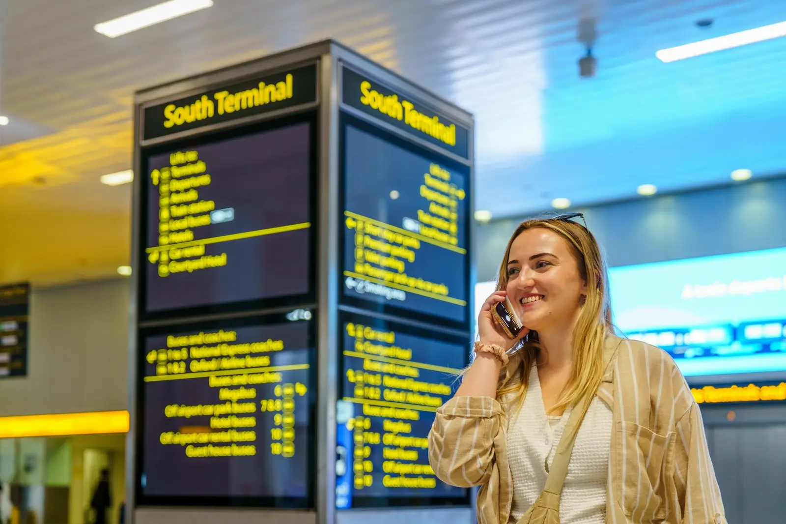 A woman on her phone in an airport arrivals terminal in front of a large information board